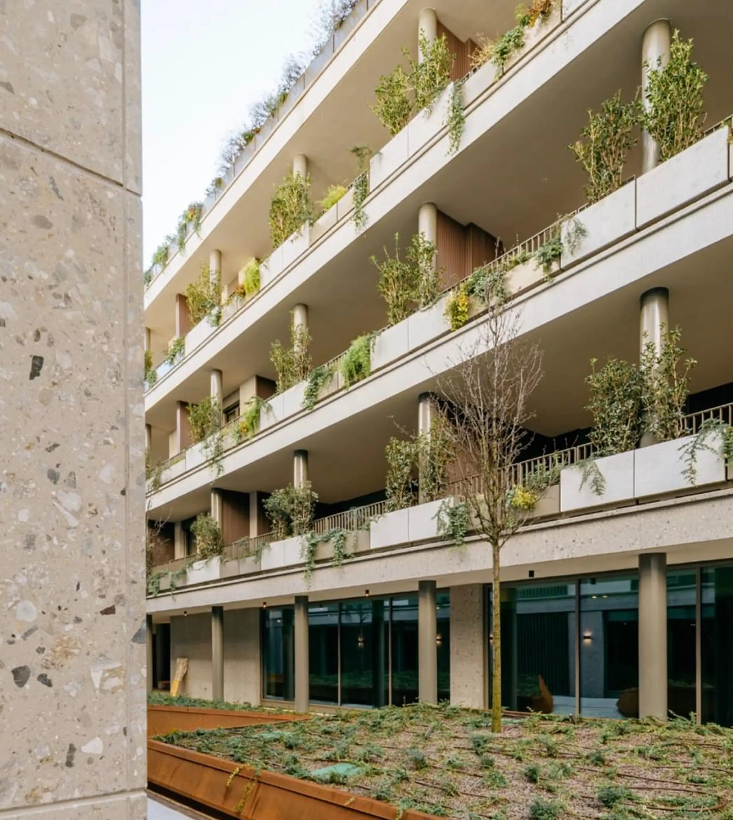 Cortile interno di complesso residenziale di lusso con facciata in pietra naturale e colonna in Travertino in primo piano, balconi a verde pensile su più livelli e hall con vetrate a piano terra – lavorazione e posa in opera facciate di pregio, Trapattoni Marmi