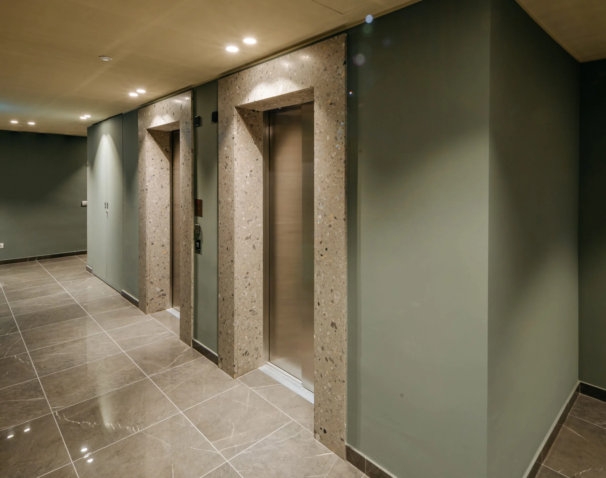 Corridoio ascensori del complesso Adamello Living Romana Yards con contorni in Agglotech SB130 Aggloceppo levigato, pavimento in marmo grigio e pareti in tono verde salvia - posa in opera Trapattoni Marmi, Milano