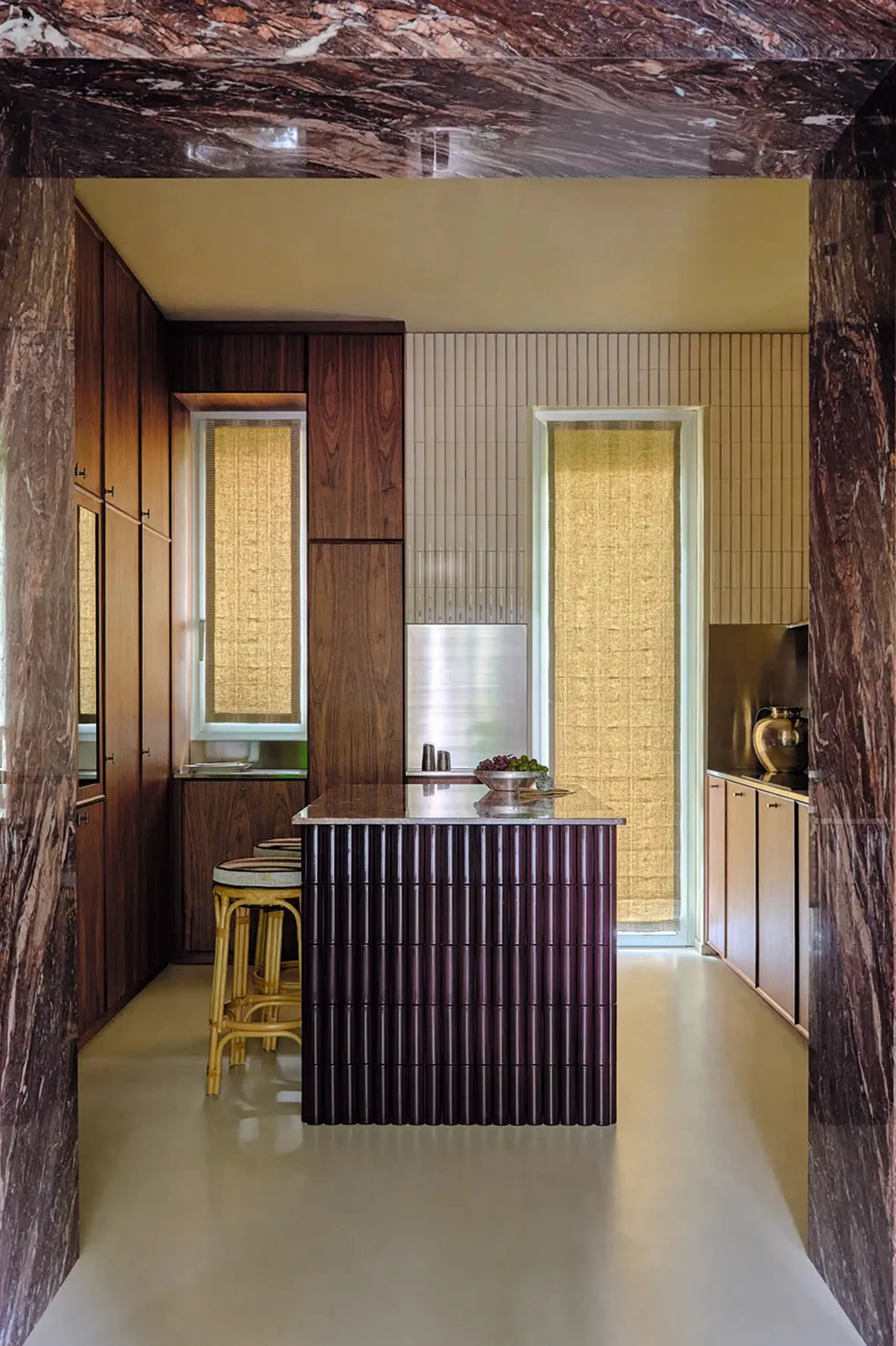 Vista della cucina attraverso il portale d'ingresso in Cipollino rosso-viola lucido: isola con piano in marmo, mobili in noce Canaletto, piano cottura a induzione e piastrelle verticali crema - dimora privata ai Navigli di Milano, LC Atelier x Trapattoni Marmi